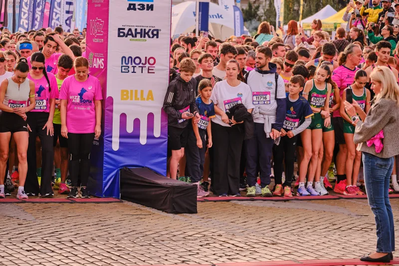 Runners lined up beside a tall pink event banner on the cobbled square.
