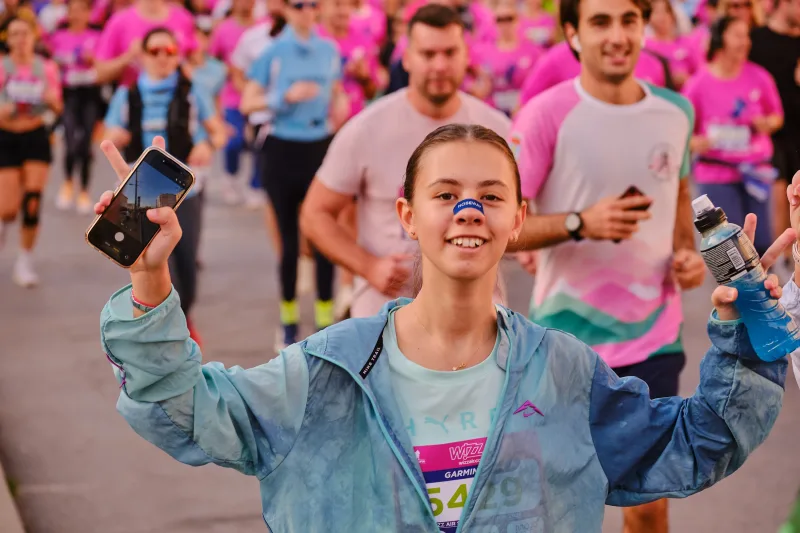 A smiling runner in a light blue jacket raising her arms with a phone in hand.