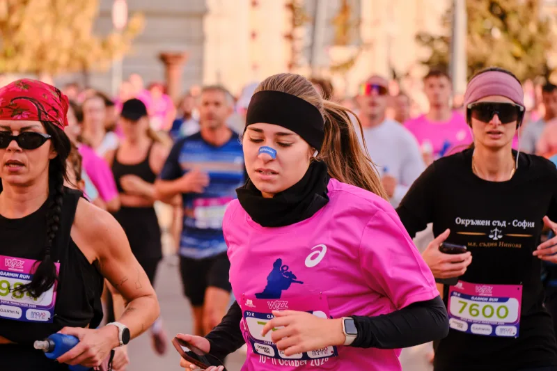 A woman in a pink shirt, black neck warmer and earphones running.