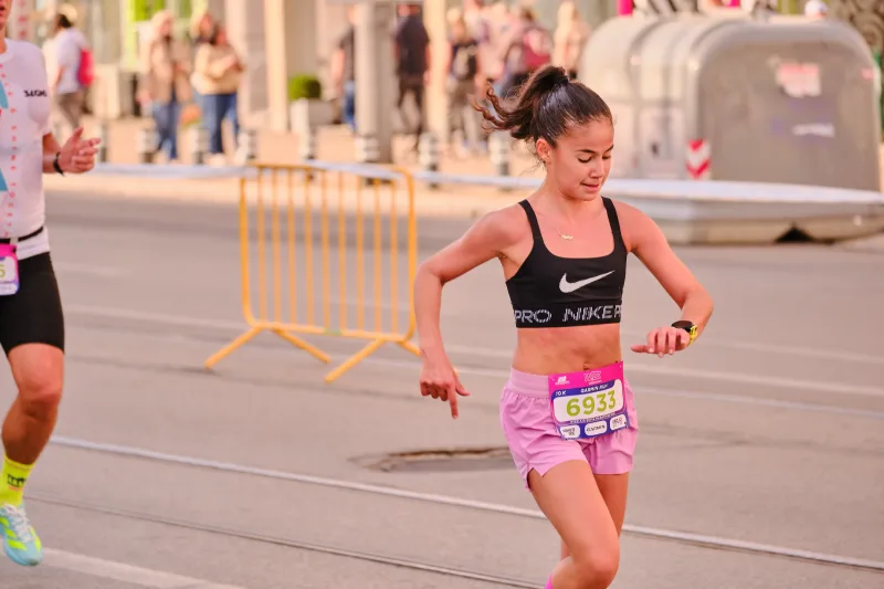 A runner in a dark sports top and pink shorts looking at her wrist.