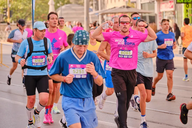 An older runner in blue leading a group through the sunlit street.