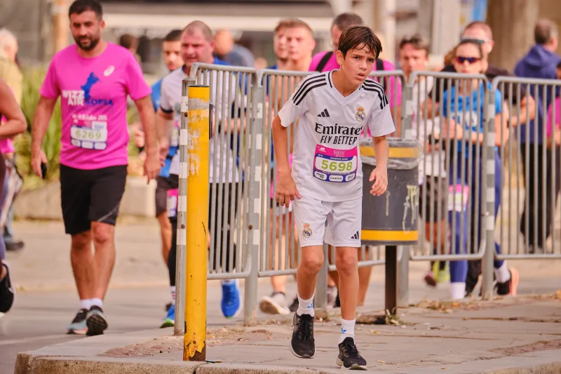 A young participant in a white football kit beside the race barriers.